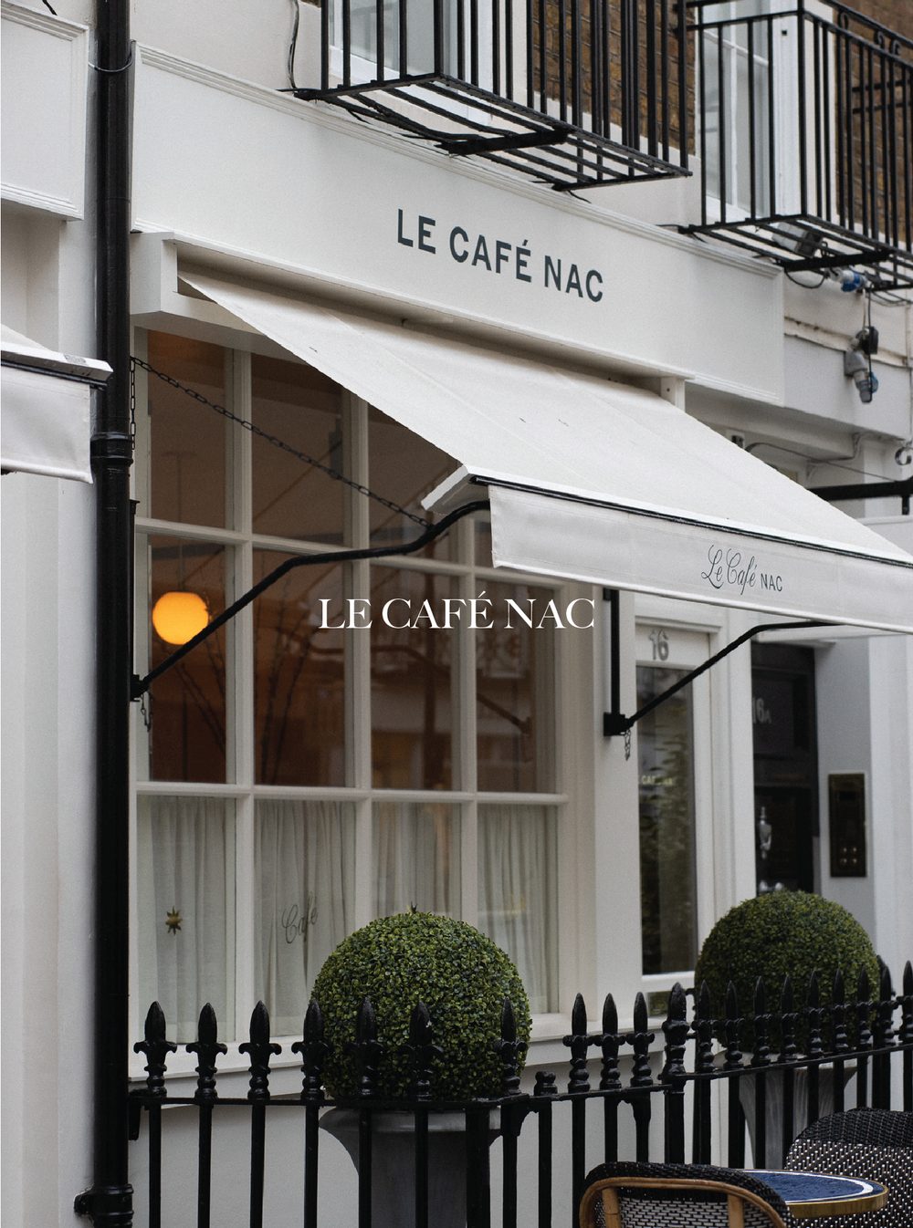 The exterior of Le Café NAC with a white storefront, black wrought iron railing fencing, potted topiary bushes, a window with white curtains, and a small round table with a yellow trim and wicker chairs outside. Le Cafe NAC.