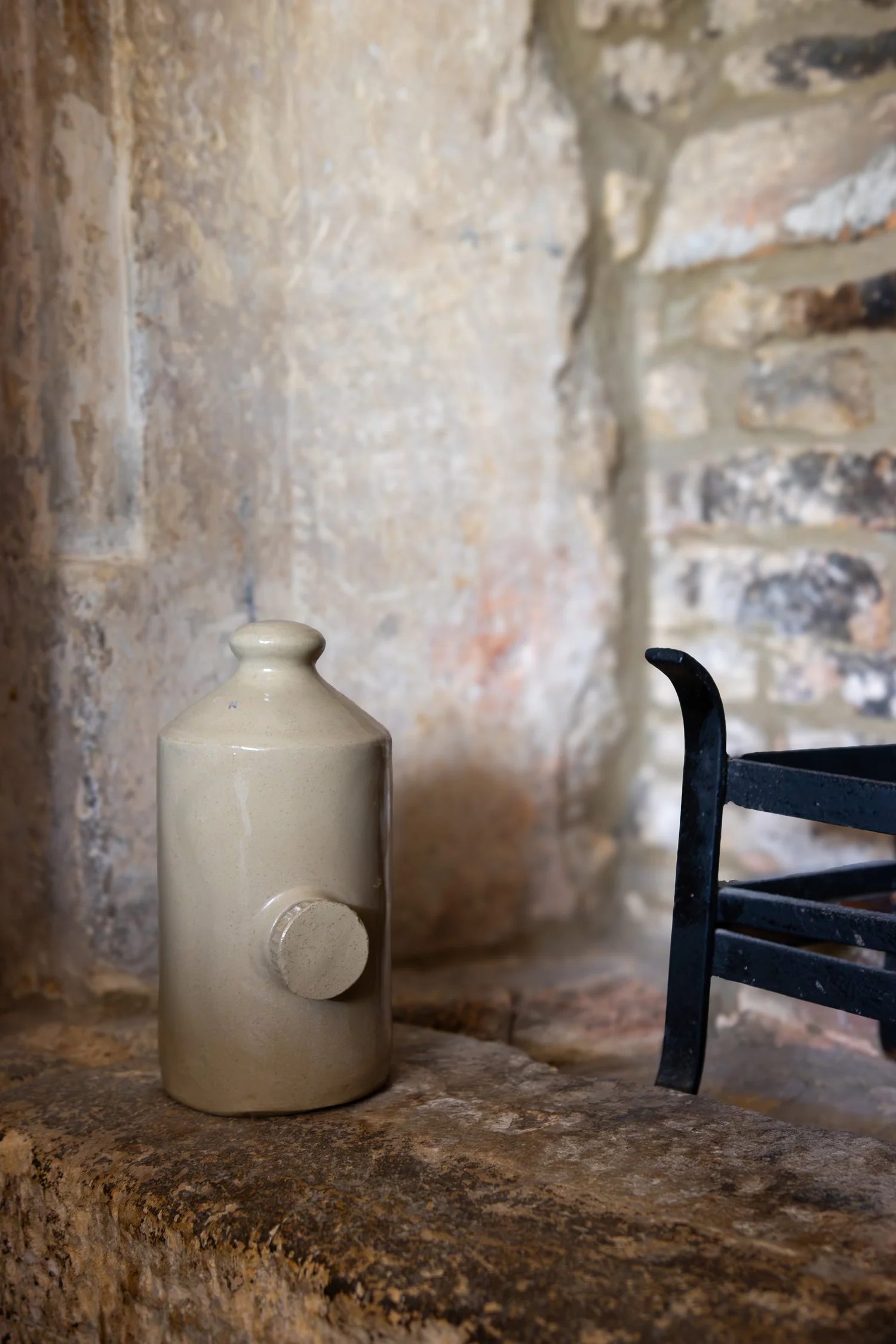 A beige ceramic bottle with a round stopper placed on a rough stone surface, in front of a brick wall background with a black metal curtain rod or pipe to the right. The Lamb Inn. 