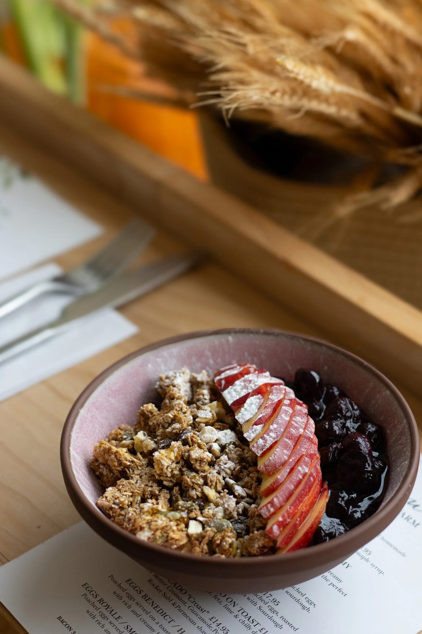 A bowl of yogurt topped with granola, sliced strawberries, and blueberries, placed on a menu or paper on a wooden table. The Cotswold Farm. 