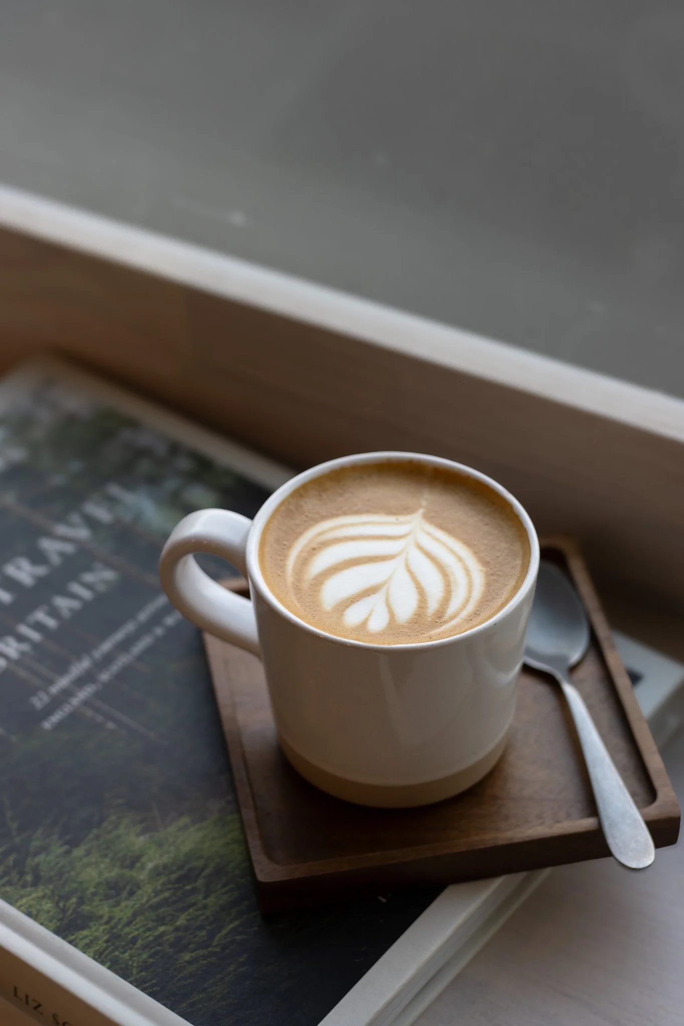 A white mug of coffee with latte art sitting on a wooden tray on a table near a window. The Cotswold Farm. 