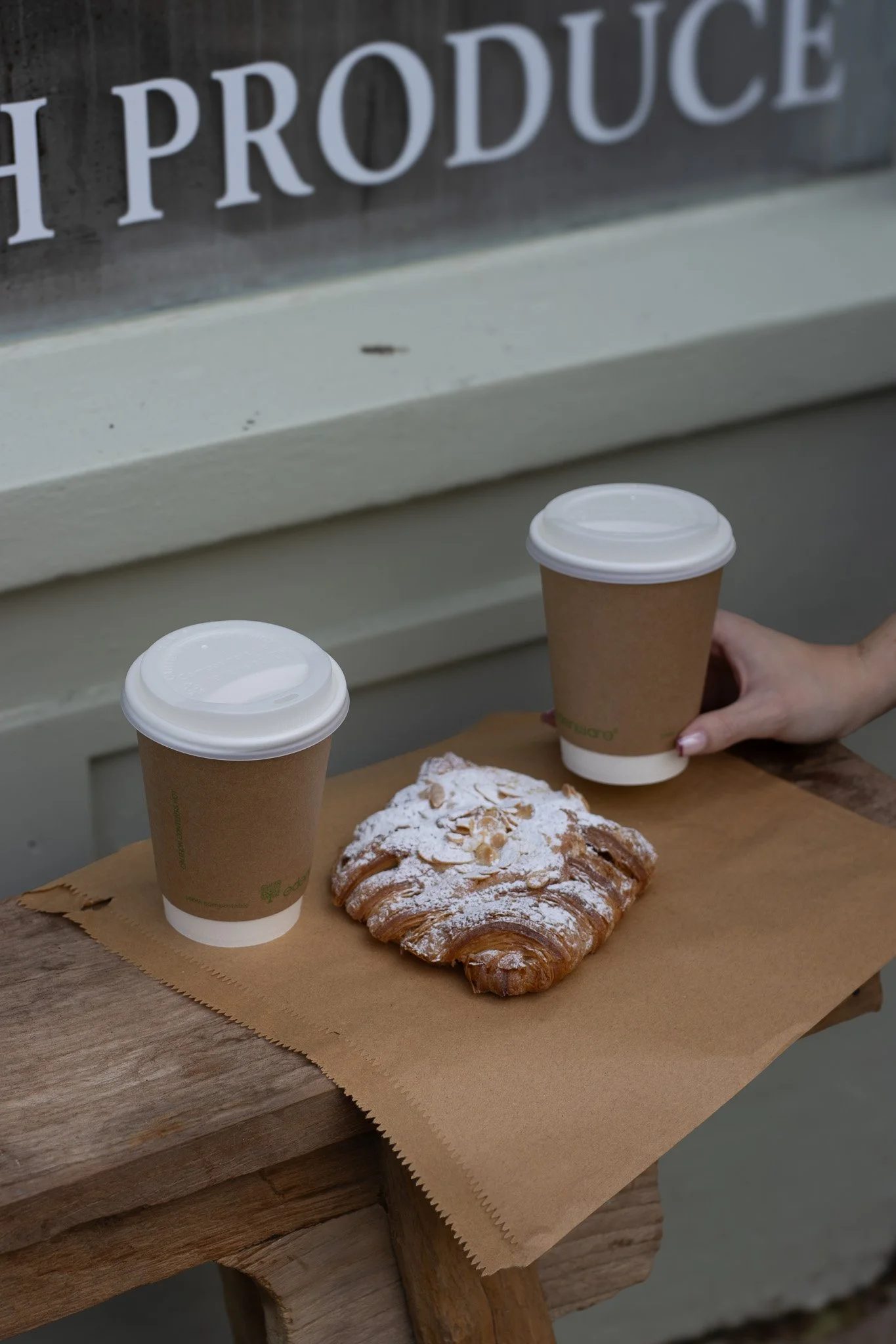Two disposable coffee cups with white lids on a wooden table, with a pastry dusted with powdered sugar and sliced almonds in front of them, and a blurry sign in the background reading 'The Produce'. The Cotswold Farm. 