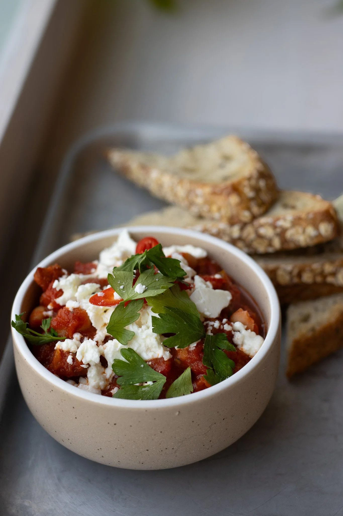 A bowl of tomato-based dish topped with crumbled cheese and fresh parsley, with bread in the background. The Cotswold Farm. 