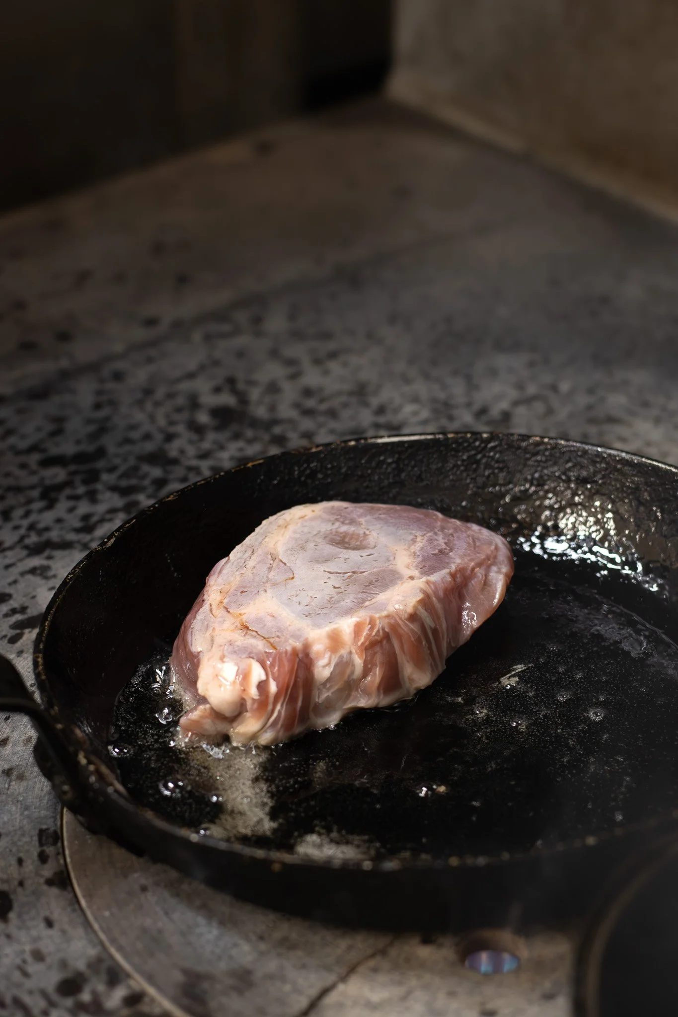 A piece of raw meat being cooked in a black cast iron skillet on a stovetop. The Lamb Inn. 