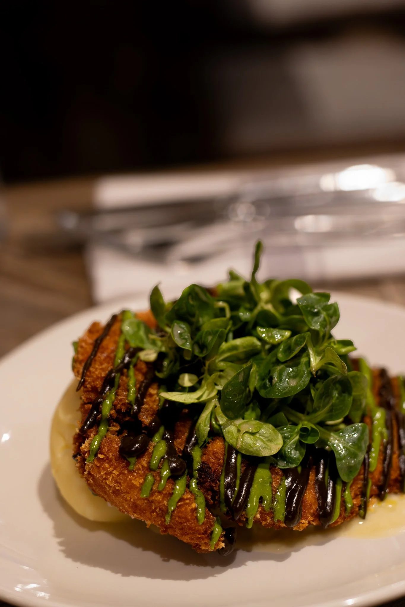 A plate of food featuring a breaded and fried meat cutlet topped with fresh green leafy salad and a drizzle of dark and green sauce, with a soft-boiled egg underneath, and a blurred background of kitchen appliances. The Lamb Inn. 