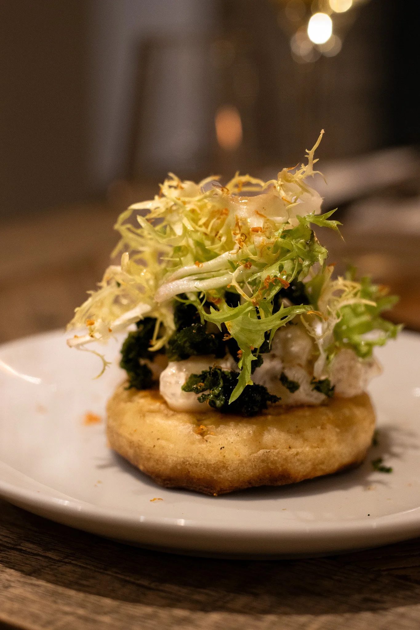 A small gourmet appetizer featuring a toasted round piece of bread topped with goat cheese, wilted greens, and microgreens, served on a white plate. The Lamb Inn. 