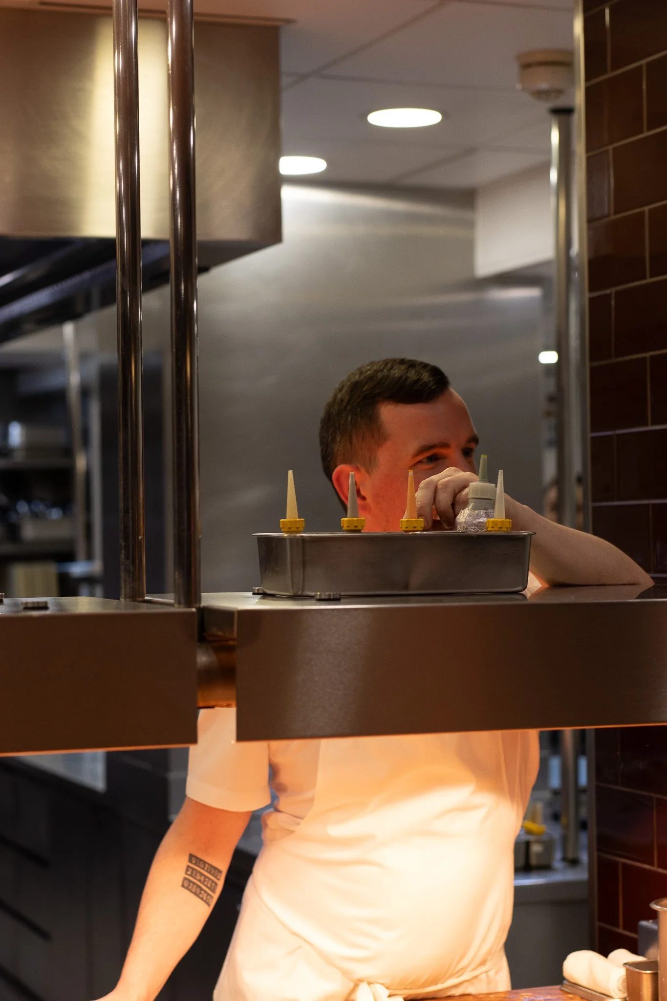 A person with short hair and a tattoo on their left arm, wearing a white shirt, is working behind a counter in a restaurant or kitchen. The background shows a commercial kitchen setting with stainless steel. ORSON VERGNAUD. Petrus.