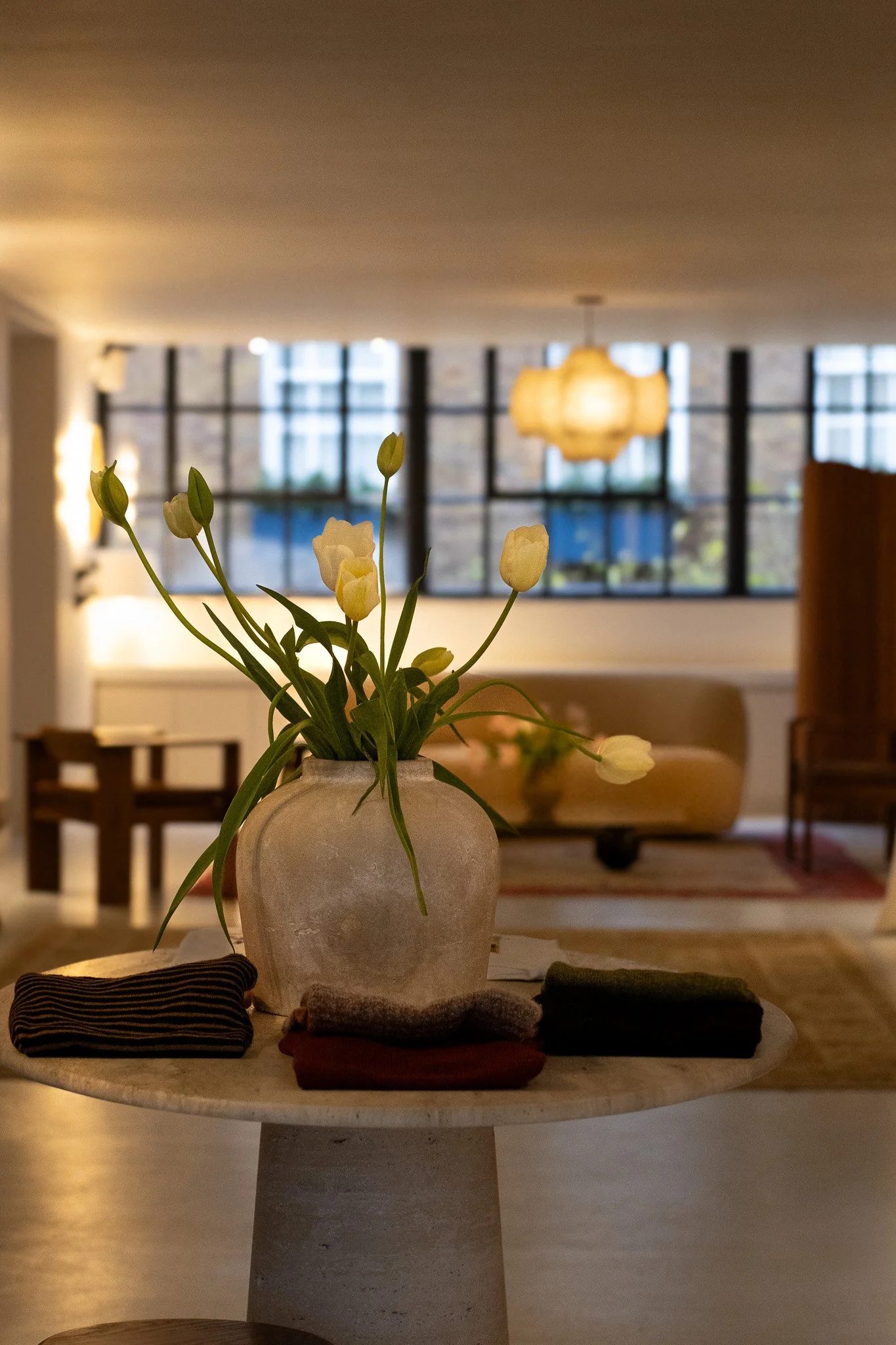 A white ceramic vase holding white tulips with unopened buds is on a round table. In the background, there is a living room with large windows, a sofa, a wooden chair, a hanging light fixture, and a cozy, well-lit interior. Realisation Par.