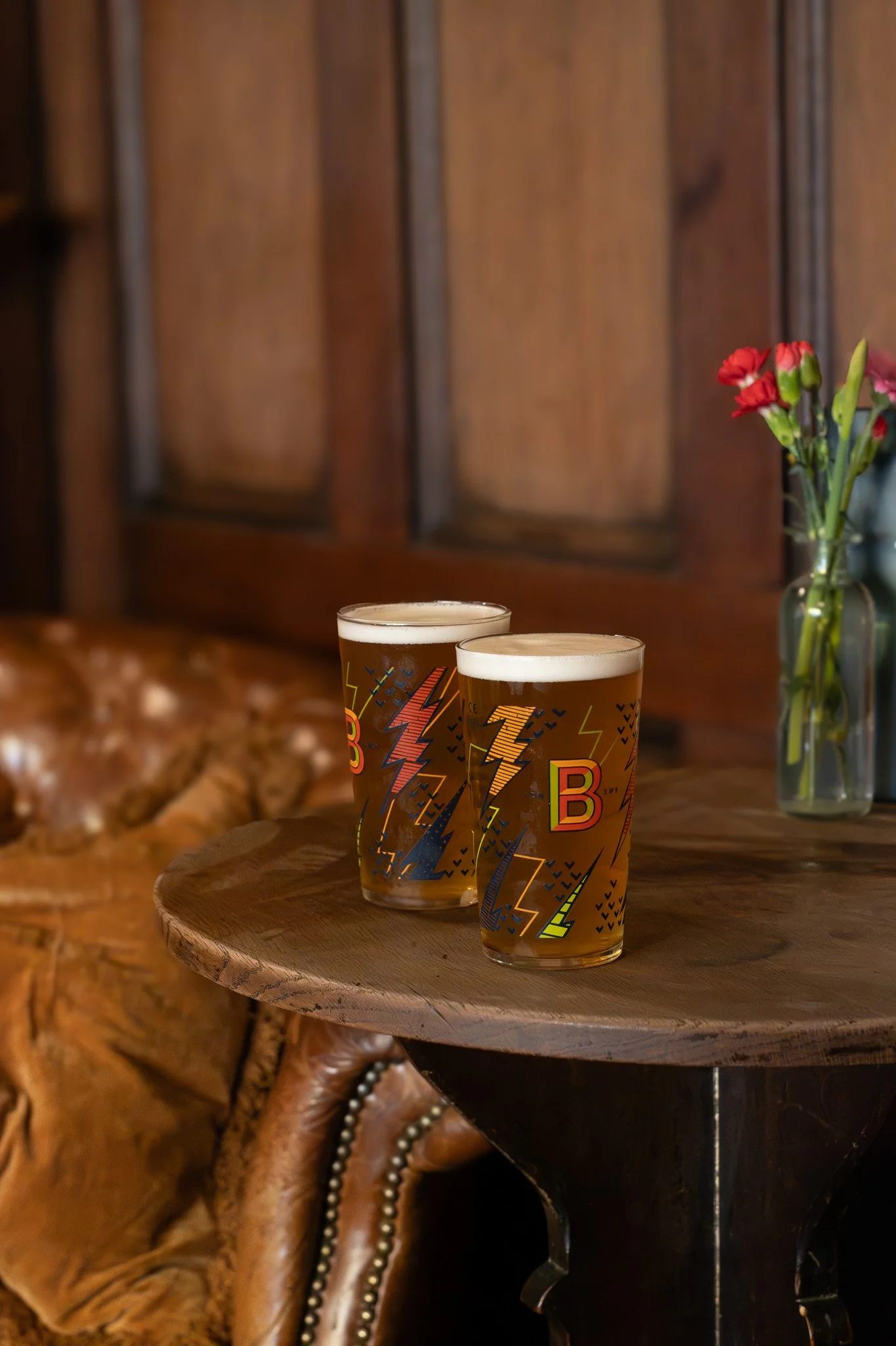 Two glasses of beer on a wooden table with a leather chair and a vase of red flowers in the background.  The Wilton Arms.