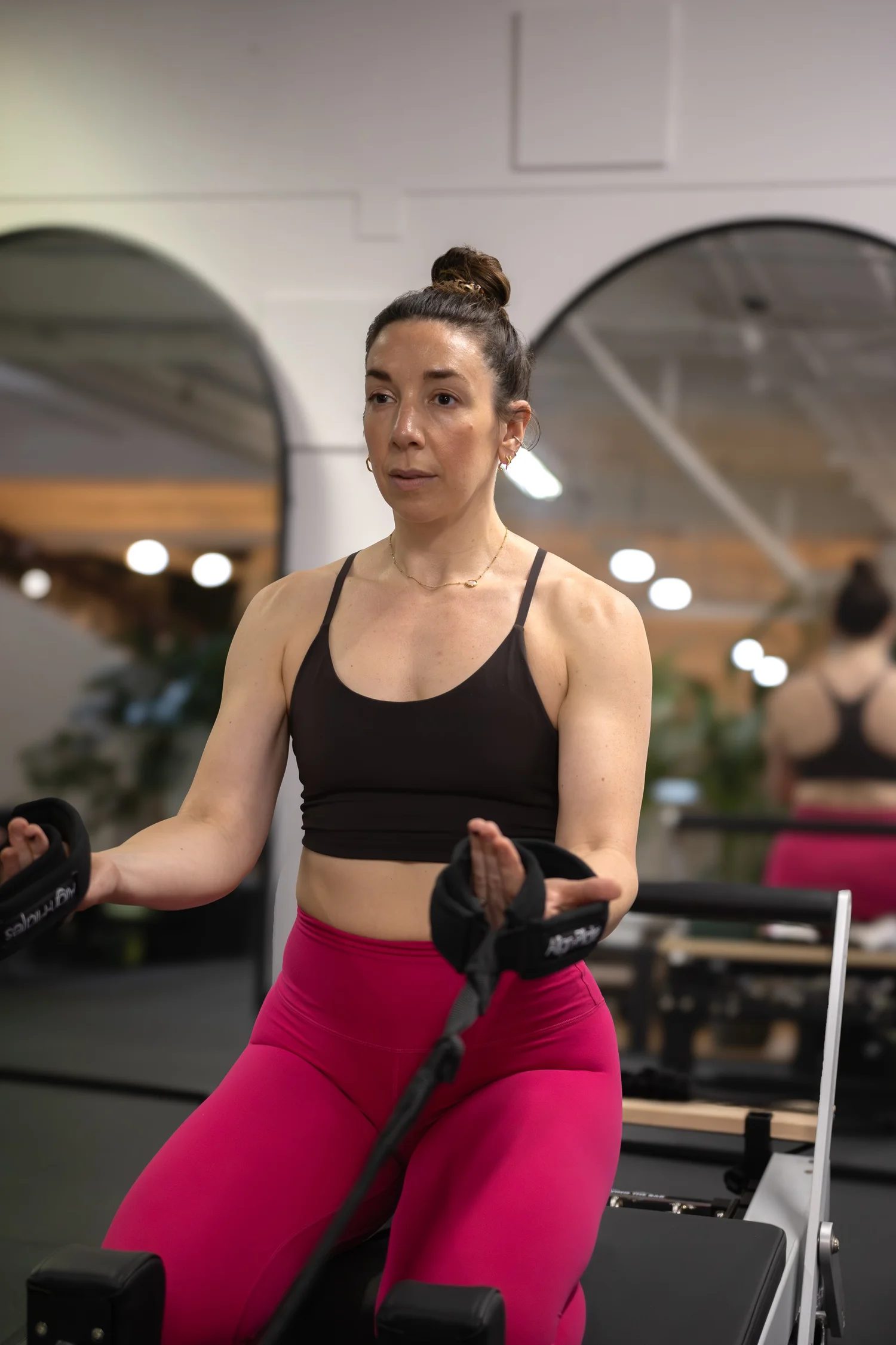 A woman with dark hair tied in a bun, wearing a black sports bra and bright pink leggings, is exercising on a rowing machine in a gym. The background shows mirrors and gym lighting. The Studio Cheltenham, Reformer Pilates. 