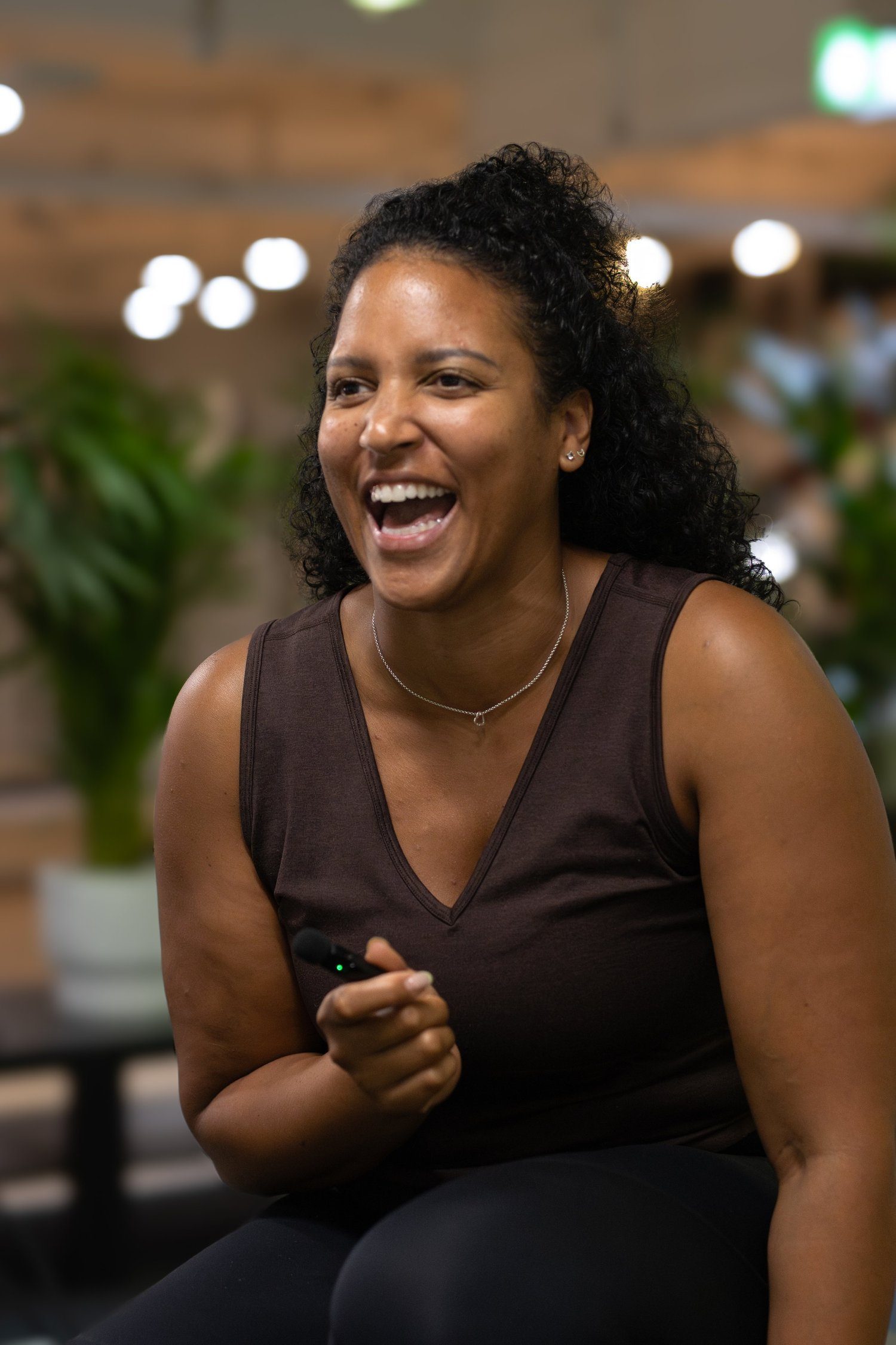 A woman smiling and holding a remote control in her hand, sitting indoors with blurred lights and plants in the background. The Studio Cheltenham, Reformer Pilates. 